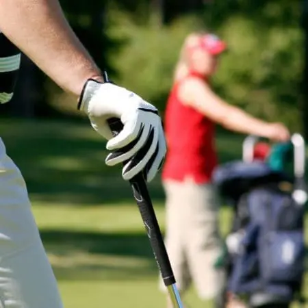 Golfurlaub in Vorarlberg, Montafon Nahaufnahme der behandschuhten Hand eines Golfspielers, der einen Schläger greift, mit einem unscharfen Hintergrund, der eine andere Person in einem roten Oberteil und einer Mütze zeigt, die einen Golfwagen schiebt - so wird die entspannte Atmosphäre eines Golfurlaubs auf einem üppigen, grünen Platz eingefangen.
