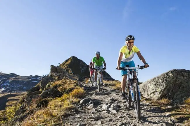 Bikeurlaub im Montafon, Vorarlberg Drei Personen fahren mit dem Mountainbike auf einem felsigen Weg, der von grasbewachsenen Hügeln und Felsen umgeben ist, unter einem klaren blauen Himmel. Der Anführer, der die Annehmlichkeiten der freien Natur genießt, lächelt und trägt einen Helm, kurze Hosen und ein Radtrikot.