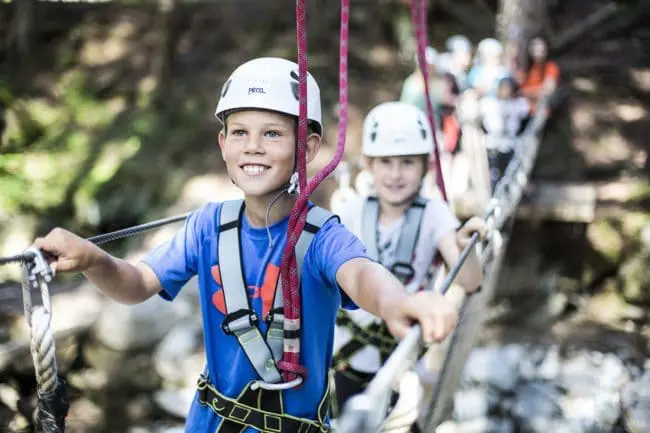 Familienurlaub im Montafon, Vorarlberg Two children wearing helmets and harnesses cross a rope bridge in a forest, smiling and holding onto the cables—perfect for family holidays. Several people are blurred in the background, also enjoying the bright outdoor adventure.
