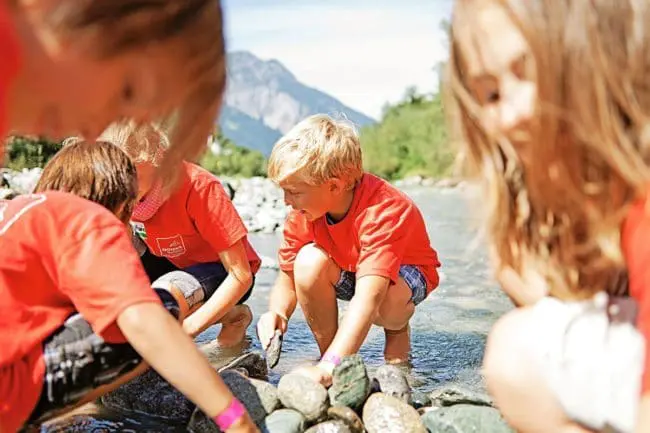 Familienurlaub im Montafon, Vorarlberg Four children wearing red shirts crouch by a shallow river, picking up rocks and playing in the water during family holidays, with mountains and greenery visible in the background on a sunny day.