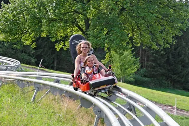 Familienurlaub im Montafon, Vorarlberg A woman and a young girl ride together in a red alpine coaster cart on a metal track outdoors, surrounded by green grass and trees, both looking ahead with excitement—perfect fun for family holidays.