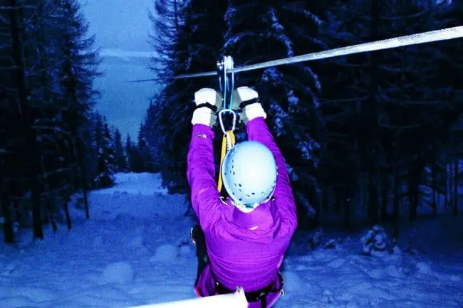Flying Fox - Winterurlaub im Montafon A person wearing a blue helmet and purple jacket rides a zipline through a snowy forest at dusk, enjoying the thrill of the winter holidays with trees and snow-covered ground visible below.