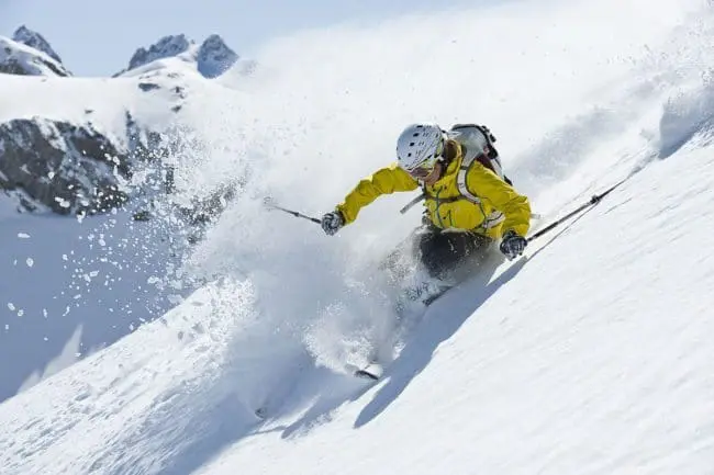 Freeride - Winterurlaub im Montafon A skier in a yellow jacket and white helmet carves down a steep, snowy slope during winter holidays, sending up a spray of powder against a backdrop of snow-covered mountains under a clear blue sky.