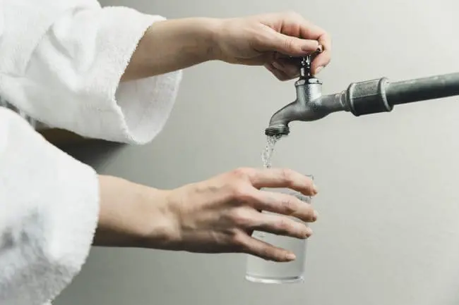 Sauberes Quellwasser - Alpenhotel Montafon, Schruns Eine Person in einem weißen Gewand genießt moderne Annehmlichkeiten, indem sie ein Glas mit Wasser aus einem Metallhahn füllt - mit einer Hand hält sie das Glas unter den laufenden Wasserhahn und mit der anderen dreht sie den Hahn auf.