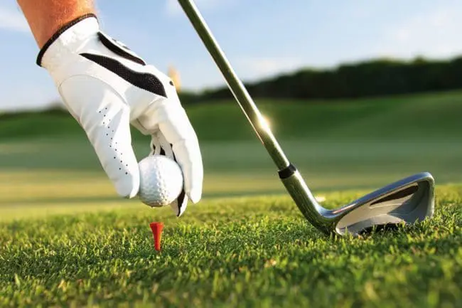 Golfurlaub im Montafon, Vorarlberg Eine behandschuhte Hand platziert einen Golfball auf einem roten Abschlag, neben einem Golfschläger auf grünem Gras, wobei die Annehmlichkeiten des Platzes im Hintergrund leicht verschwimmen.