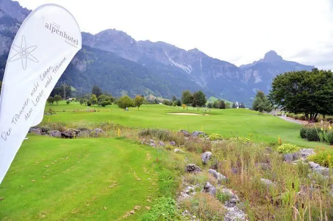 Golfplatz Montafon, Vorarlberg Ein malerischer Golfplatz mit gepflegtem grünem Rasen, perfekt für einen Golfurlaub, ist umgeben von Bergen und Bäumen unter einem bewölkten Himmel. Ein weißes Alpenhotel-Banner steht auf der linken Seite neben Wildblumen und Felsen.
