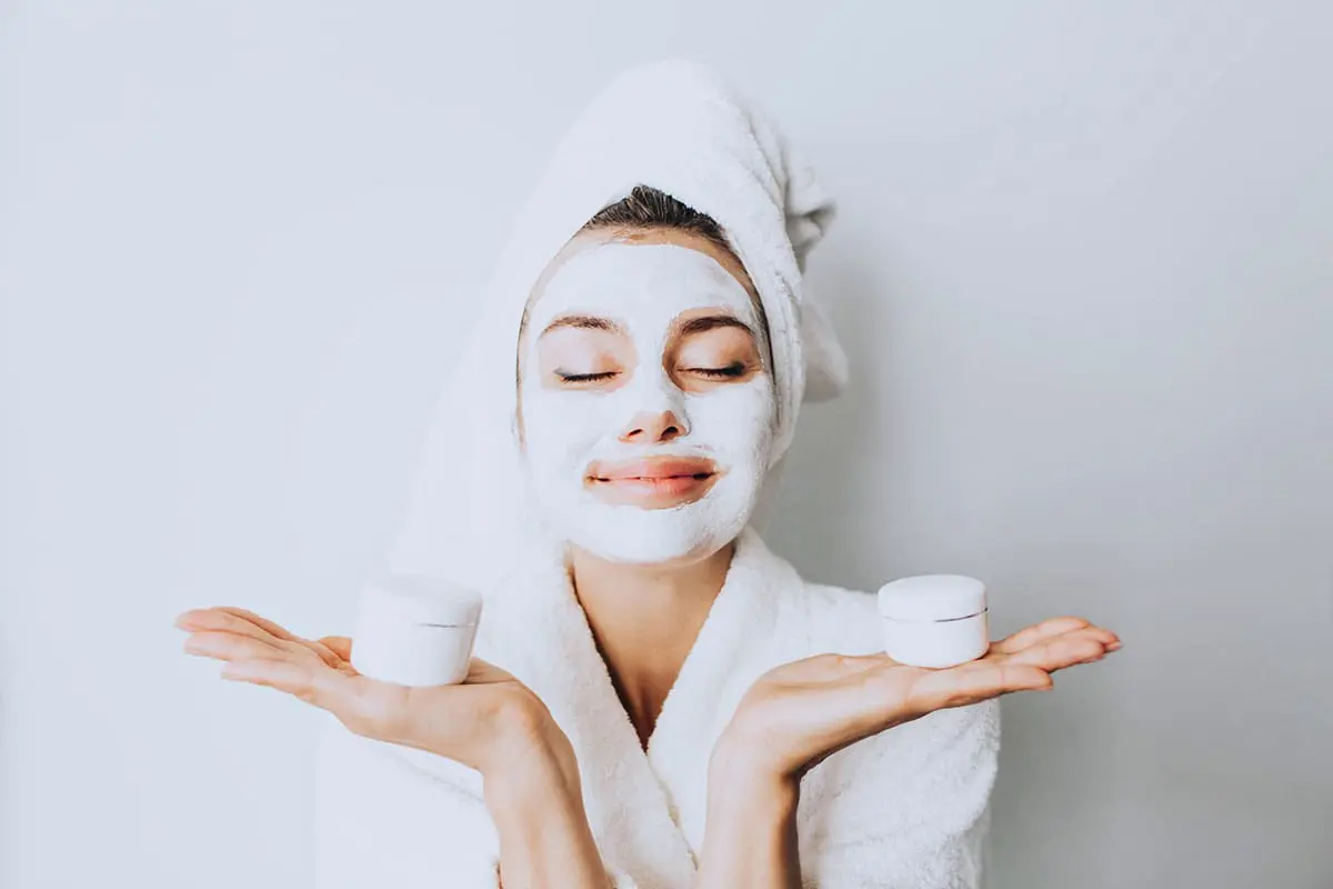 Eine Frau in einem weißen Bademantel und einem Handtuch wickelt sich die Haare, lächelt mit geschlossenen Augen und trägt eine Gesichtsmaske. Sie hält zwei kleine weiße Kosmetiktiegel, einen in jeder Hand, vor einem schlichten hellen Hintergrund.