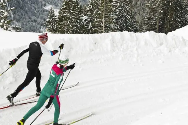 Langlaufen - Winterurlaub im Montafon Two people cross-country skiing on a snowy trail surrounded by trees and snowbanks, with mountains in the background—an ideal scene for winter holidays. Both are wearing winter sports clothing and hats.