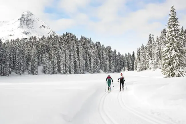 Langlaufen - Winterurlaub im Montafon Two people cross-country skiing on a snowy trail through a winter forest, enjoying the serene beauty of tall snow-covered trees and a mountain peak under a partly cloudy sky during their winter holidays.