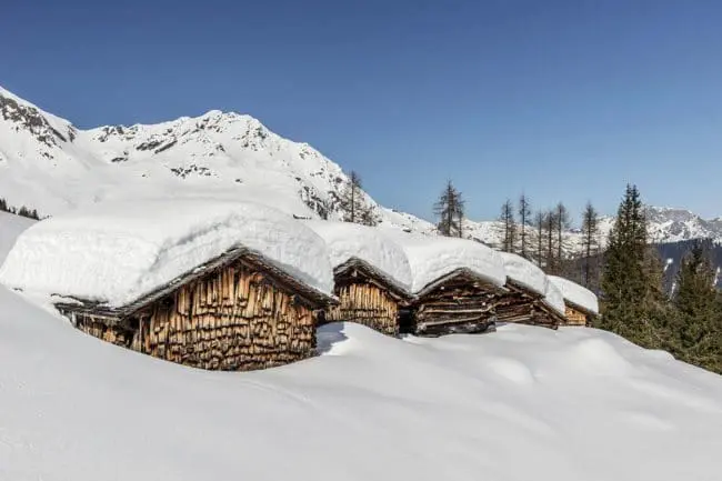 Montafon, Vorarlberg - Fotografie Manfred Schlatter Vier Holzhütten mit schneebedeckten Dächern sind eingebettet in eine verschneite Berglandschaft, umgeben von immergrünen Bäumen und schneebedeckten Gipfeln unter einem klaren blauen Himmel, und bieten einen ruhigen Rückzugsort, der die alpine Kultur zelebriert.