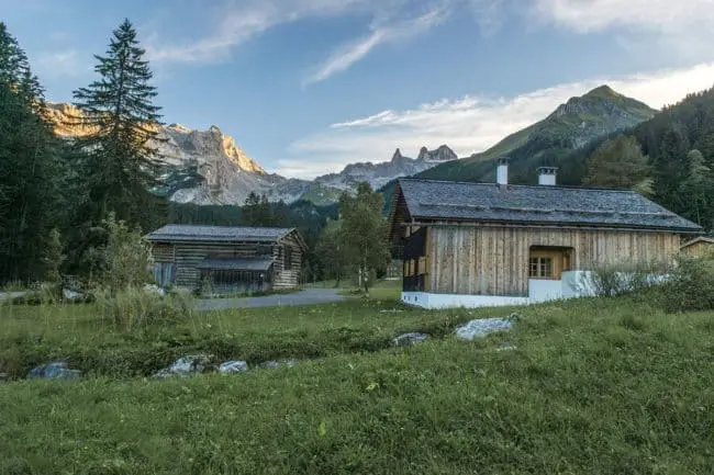 Montafon, Vorarlberg - Fotografie Manfred Schlatter Zwei rustikale Holzhütten stehen auf einer von Bäumen umgebenen Wiese, wo sich der Geist der lokalen Kultur mit der Natur und den zerklüfteten Bergen vermischt, die sich im Hintergrund unter einem teilweise bewölkten Himmel bei Sonnenuntergang erheben.