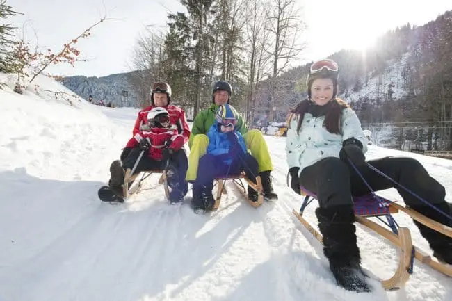 Rodeln - Winterurlaub im Montafon Four people in winter clothes sit on sleds in the snow, smiling at the camera during their winter holidays. Trees and mountains are visible in the background under a bright sun.
