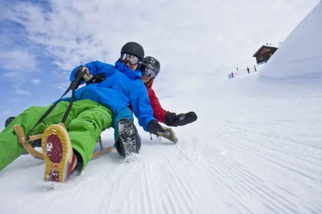 Rodeln - Winterurlaub im Montafon Two people wearing helmets and winter clothing sled down a snowy hill at high speed, enjoying the thrill of winter holidays, with a wooden building and others in the background under a partly cloudy sky.