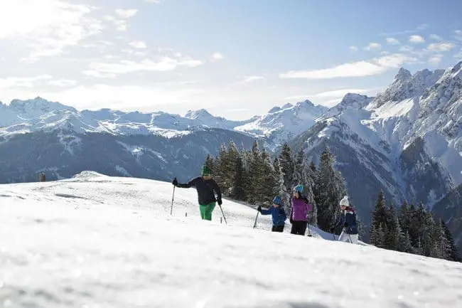 Schneeschuhwandern - Winterurlaub im Montafon A group of people trek across a snowy mountain slope with walking poles, surrounded by evergreen trees and distant snow-covered peaks—a perfect scene for winter holidays under a partly cloudy sky.