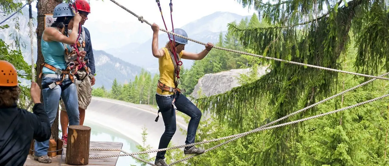Seminare & Incentives im Alpenhotel Montafon, Vorarlberg Vier Personen mit Helmen und Klettergurten überqueren einen Hochseilgarten in der Nähe eines Luxushotels in einer bewaldeten, bergigen Gegend. Eine Person geht vorsichtig zwischen den Seilen hindurch, während andere warten und helfen, umgeben von üppig grünen Bäumen und dem Blick auf die Berge in der Ferne.