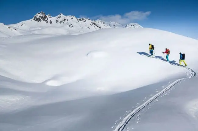 Skitouren - Winterurlaub im Montafon Three people in colorful winter gear hike up a snow-covered mountain slope, leaving a trail behind them—an adventurous way to spend the winter holidays, surrounded by snowy peaks and a clear blue sky.