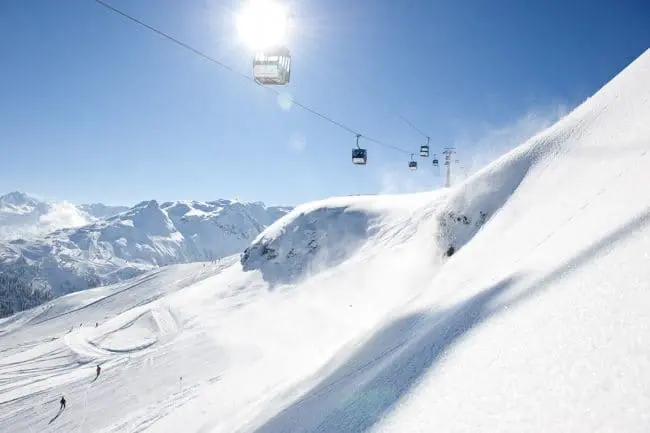 Skiurlaub im Montafon, Vorarlberg Ski lifts move above snowy slopes and mountains under a bright sun and clear blue sky, capturing the magic of winter holidays. Skiers are visible in the distance, with powdery snow blowing on the hillside.