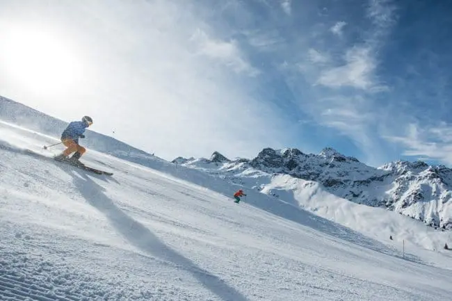 Skiurlaub im Montafon, Vorarlberg Two skiers descend a snowy mountain slope under a bright sun and blue sky, enjoying unforgettable winter holidays with rugged, snow-covered peaks in the background.