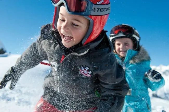 Skiurlaub im Montafon, Vorarlberg Two children wearing ski gear and helmets play joyfully in the snow, laughing as snow sprays around them under a clear blue sky, capturing the magic of winter holidays.