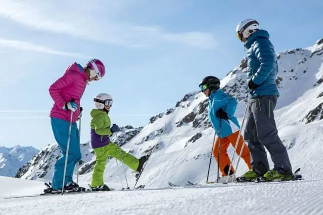 Skiurlaub im Montafon, Vorarlberg A family of four in colorful ski outfits stands on a snowy mountain slope, preparing to ski during their winter holidays. The adults watch as a child lifts a ski, with bright blue skies and snow-covered peaks in the background.