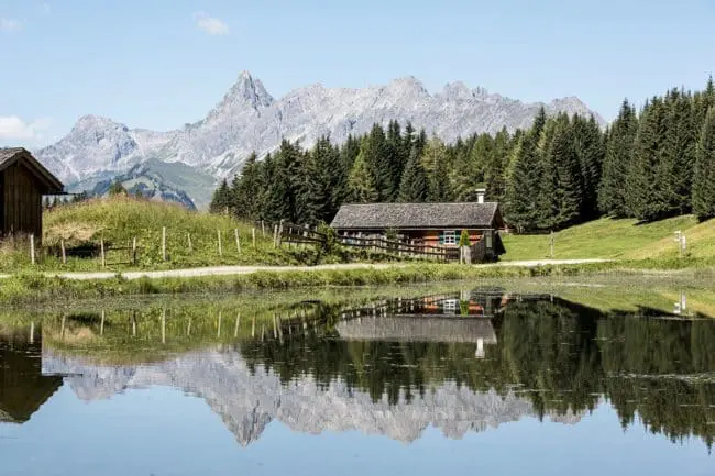 Sommerurlaub im Montafon, Vorarlberg Eine Holzhütte liegt an einem Teich, umgeben von grünem Gras und hohen Kiefern - ein idealer Ort für einen ruhigen Sommerurlaub. Die Bergketten spiegeln sich im ruhigen Wasser unter dem blauen Himmel.
