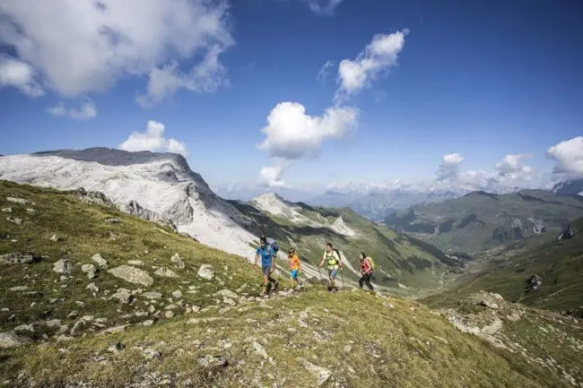 Wanderurlaub im Montafon, Vorarlberg Eine Gruppe von fünf Personen genießt das Wandern auf einem grasbewachsenen Bergpfad mit felsigen Gipfeln und einem weitläufigen Tal im Hintergrund, unter einem strahlend blauen Himmel mit vereinzelten Wolken.