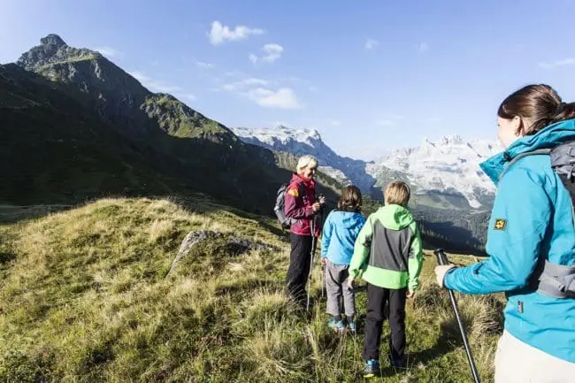 Wanderurlaub im Montafon, Vorarlberg Vier Personen in Outdoor-Kleidung und Rucksäcken stehen auf einem grasbewachsenen Hügel und blicken auf Berge unter einem blauen Himmel mit vereinzelten Wolken. Die Gruppe scheint beim Wandern in einer malerischen Alpenlandschaft zu sein.