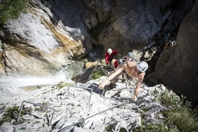 Wanderurlaub im Montafon, Vorarlberg Drei Personen mit Helmen klettern über Metallsprossen und Sicherungsseile auf einem Klettersteig eine steile Felswand hinauf, während eine weitere Person in der Nähe des Fußes wartet. Die Szene zeigt das abenteuerliche Wandern mit felsigem Gelände und einem Bach unterhalb.