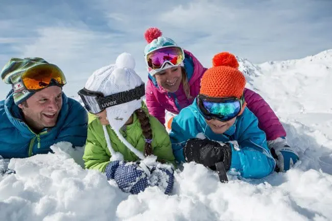 Winterwandern - Winterurlaub im Montafon A family of four wearing colorful winter clothes and ski goggles lies in the snow, smiling and enjoying a sunny day surrounded by snowy mountains during their winter holidays.